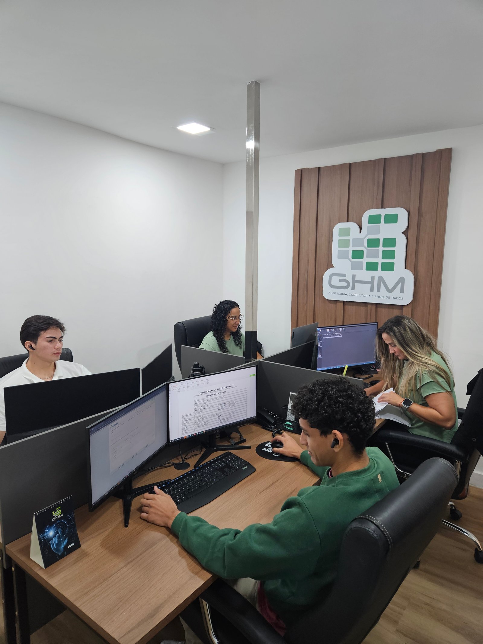 Equipe GHM em atendimento no escritório, com painel e logotipo da empresa ao fundo.